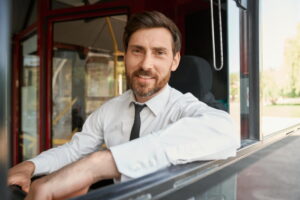 Un chauffeur de bus souriant en chemise blanche et cravate à sa fenêtre, illustrant un service de location autocar avec chauffeur.