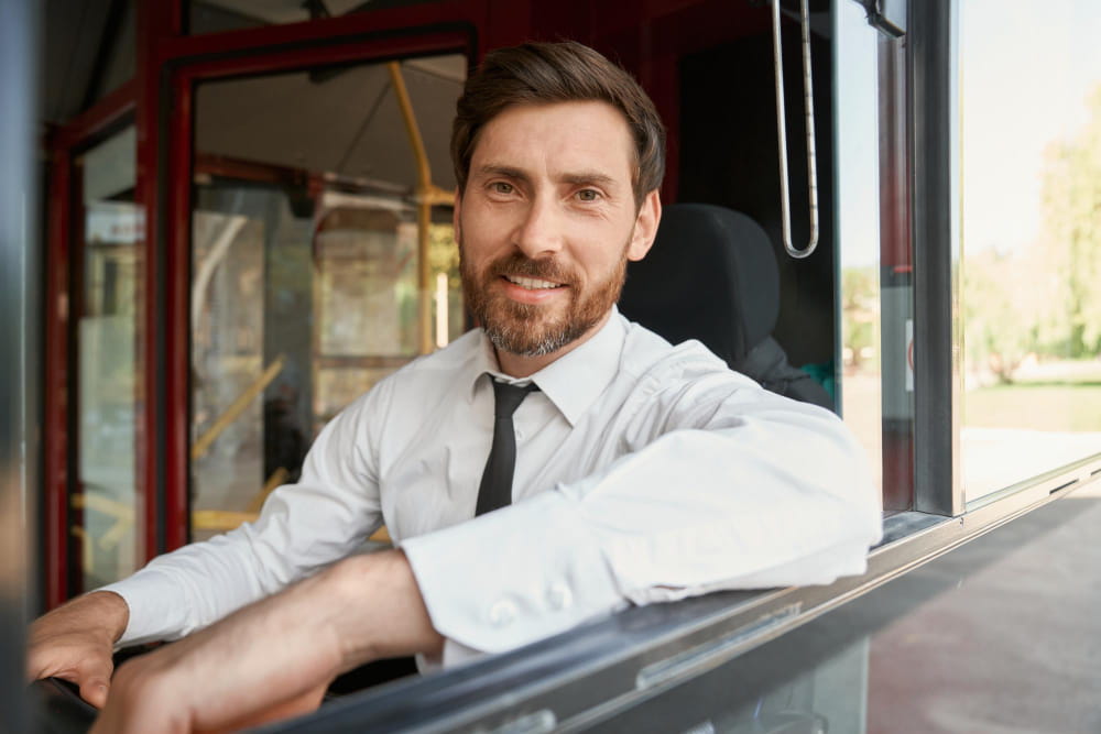 Un chauffeur de bus souriant en chemise blanche et cravate à sa fenêtre, illustrant un service de location autocar avec chauffeur.