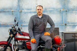 Un mécanicien en combinaison de travail grise et lunettes de sécurité est assis sur une moto chopper vintage de couleur rouge, tenant un petit outil, prêt à apprendre comment installer un pare-brise universel sur un chopper. Le décor est un garage avec un mur en béton usé.