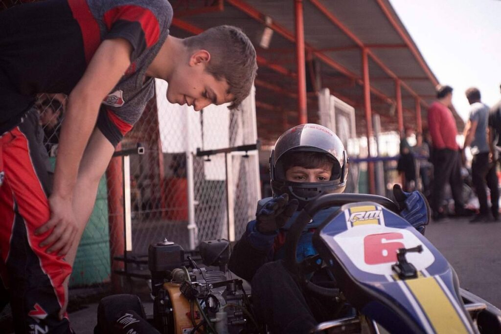 Jeune pilote en combinaison bleue et casque, assis dans un kart jaune et bleu, encadré par un adulte dans une zone de paddock