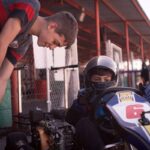 Jeune pilote en combinaison bleue et casque, assis dans un kart jaune et bleu, encadré par un adulte dans une zone de paddock