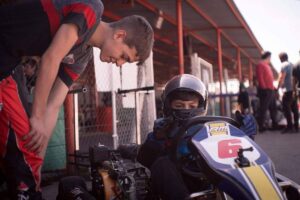 Jeune pilote en combinaison bleue et casque, assis dans un kart jaune et bleu, encadré par un adulte dans une zone de paddock