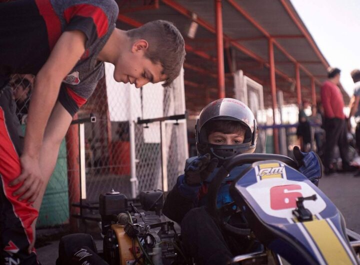 Jeune pilote en combinaison bleue et casque, assis dans un kart jaune et bleu, encadré par un adulte dans une zone de paddock